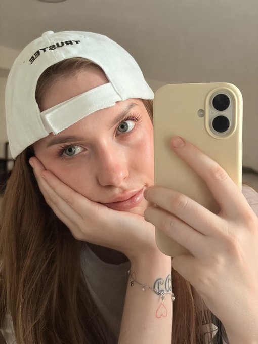 A young woman wearing a backwards white 'TSURT' cap takes a mirror selfie with a phone, resting her chin on her hands.