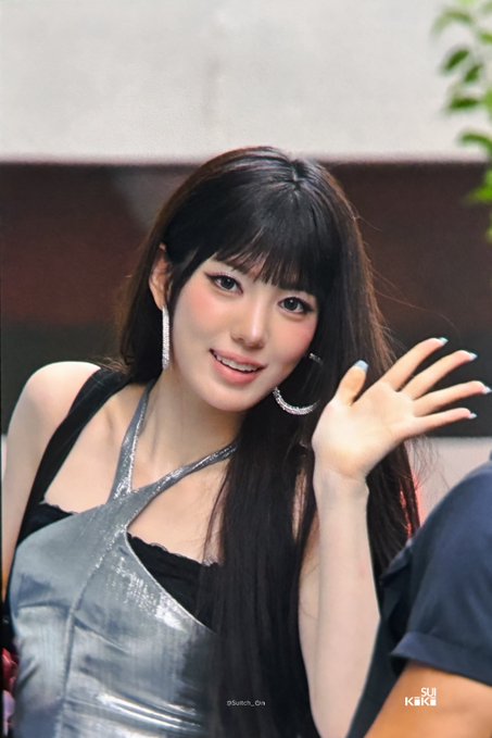 A young woman with long dark hair and bangs waving, wearing a metallic silver halter top and large hoop earrings, with a 'Sukoch_Gya' watermark.