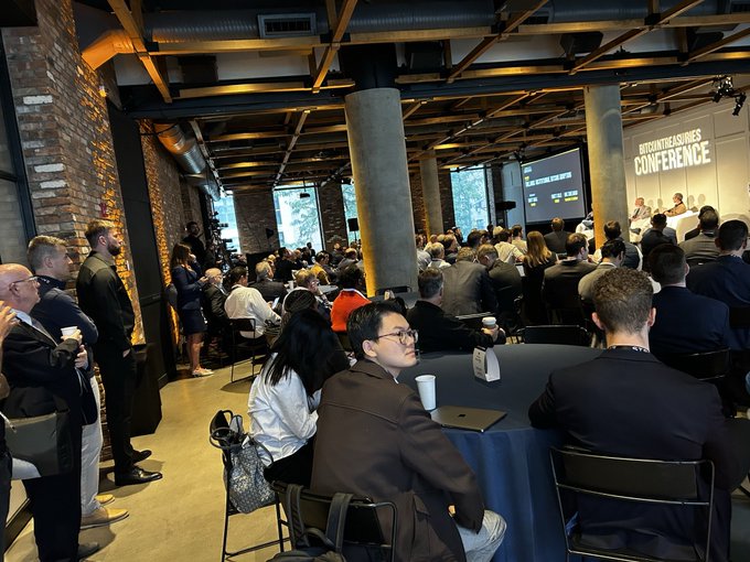 A packed audience at The Bitcoin Treasuries Conference in NYC, with attendees filling every seat at round tables and standing along the sides as presenters speak at the front, while a woman seated in the foreground captures the moment attentively. Visible on stage are banners reading "BITCOINTREASURIES" and "CONFERENCE," along with a large presentation screen, reinforcing the event's sold-out atmosphere.