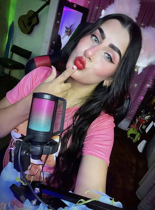 A blue-eyed woman with long black hair wearing pink fluffy ears applies lipstick while recording in front of a microphone.