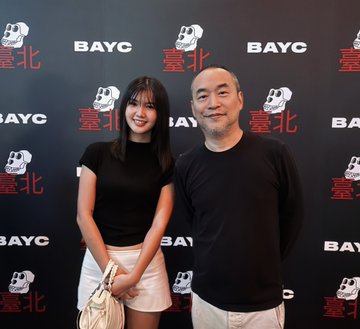 The image features a man wearing a black shirt and a woman wearing a white skirt, both smiling at the camera while posing together against a backdrop of a wall with Chinese characters for Bayc. The background includes a large "BAYC" sign that is red and white in color.