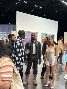 The image shows a group of people standing around, with one person wearing a white dress and another person wearing a black leather jacket. A woman in a white dress stands out among them, possibly posing for a photo or engaging in conversation. The background features an art gallery setting with paintings on the walls.
