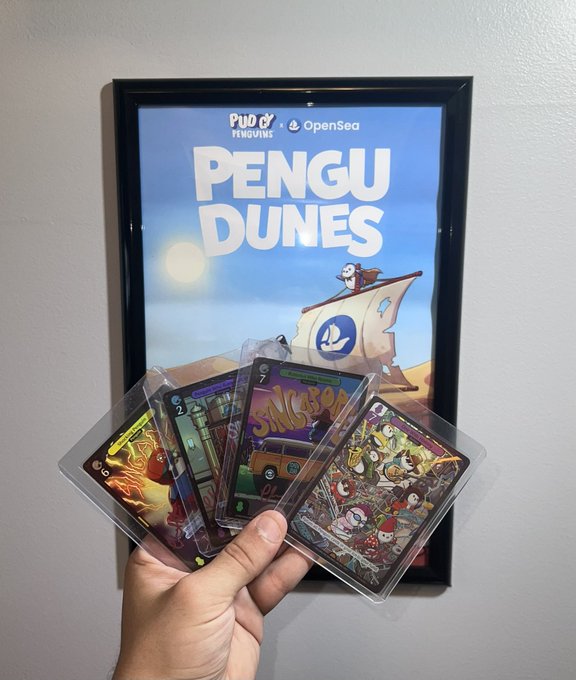 A hand holding four collectible Pudgy Penguins trading cards in front of a framed 'Pengu Dunes' poster collaboration between Pudgy Penguins and OpenSea.