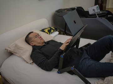A man relaxes on a bed using a portable laptop table, mirroring the casual work-from-bed style referenced in the post promoting CZ's "Laptop Table For Bed" available for 0.1 $BNB. The setup shows him comfortably propped up with the adjustable table holding his laptop, embodying the laid-back tech entrepreneur aesthetic the post alludes to.