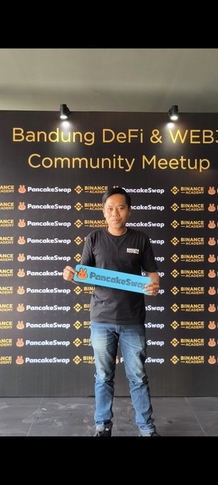 The image features a man standing against a wall with a banner that says "BANDING DEFI & WEB Community Meeting Ups" written above him. The man is holding up a blue sign, which has the words "PancakeSwap" on it. He appears to be excited about an upcoming event or gathering related to Web3 and DeFi.