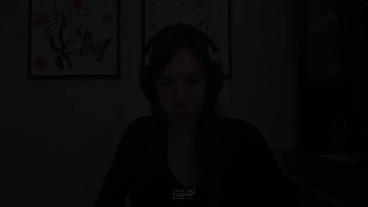 The image shows a woman wearing headphones, sitting at a desk with her laptop open in front of her. She appears to be focused on something or someone behind her, possibly a computer screen displaying an image or video. The photo has been edited to emphasize the woman's face and the background is intentionally blurred out.