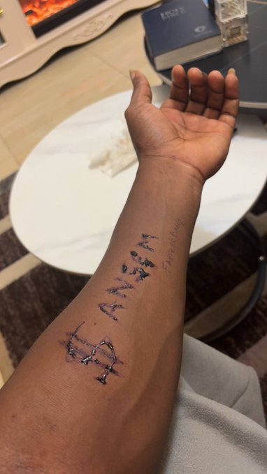 The image shows a person's forearm extended upward, featuring a freshly inked tattoo that reads "ANSEM" with "$ANSEM" written in smaller script next to it. The arm is positioned over a white round table, with what appears to be a Bible visible in the background.

The tattoo directly corresponds to the post, which mentions getting an Ansem tattoo on day 2 of "bagworking" in reference to the $ANSEM cryptocurrency/meme coin ticker.
