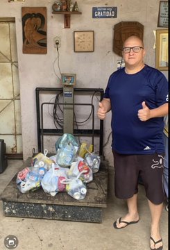 A man in a blue shirt gives two thumbs up next to a platform scale loaded with sacks of aluminum cans for recycling, symbolizing his two-year journey of collecting and selling cans to accumulate Bitcoin. Above him, a "GRATIDÃO" (gratitude) sign reflects the pride of his effort as he approaches 1 BTC.
