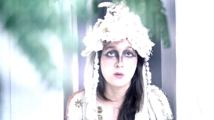 The image features a woman with white makeup, dressed in a white outfit or costume that includes a headpiece made of flowers. The background appears blurry, but there are trees visible behind her. The caption reads "NEW objkt.com/tokens/KT1Uhco". This suggests the photo might be related to an online store selling costumes and accessories, possibly for events like Halloween or themed parties.