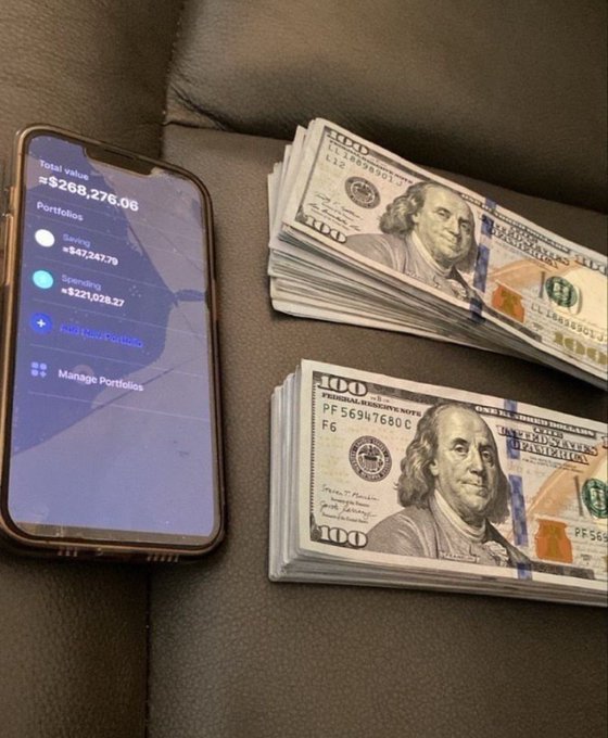 A cracked smartphone displays a Robinhood portfolio showing a total value of $268,276.06, split between a Saving portfolio of $47,247.79 and a Spending portfolio of $221,028.27, sitting next to two stacks of $100 bills—visually celebrating the "beautiful day to make money" with a brag-worthy blend of digital and physical wealth.