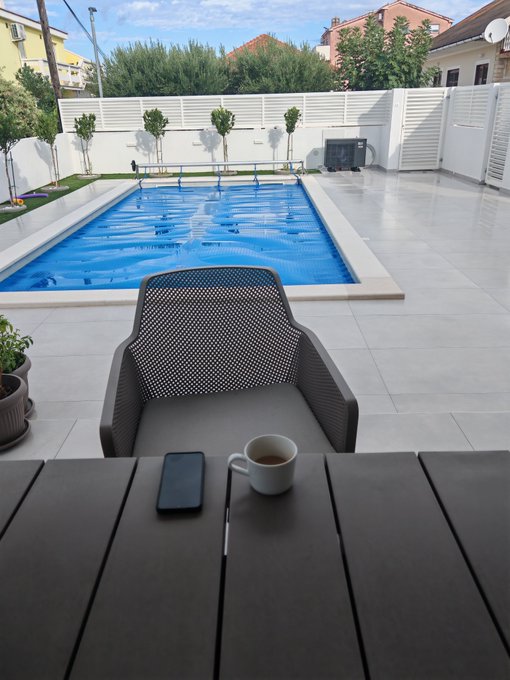 A serene backyard setting featuring a blue rectangular swimming pool, a patio chair, a table with a smartphone and a cup of coffee, all enclosed by a white fence.
