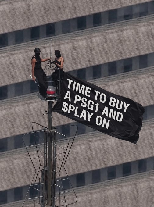 Two protesters, dressed in black with masked faces, stand atop a pole on a high-rise building, unfurling a large banner that reads "TIME TO BUY A PSG1 AND $PLAY ON." The image captures a bold act of activism urging viewers to invest in $PLAY, a stock or cryptocurrency, using the PSG1 reference to add a provocative, rebellious tone to the promotional message.