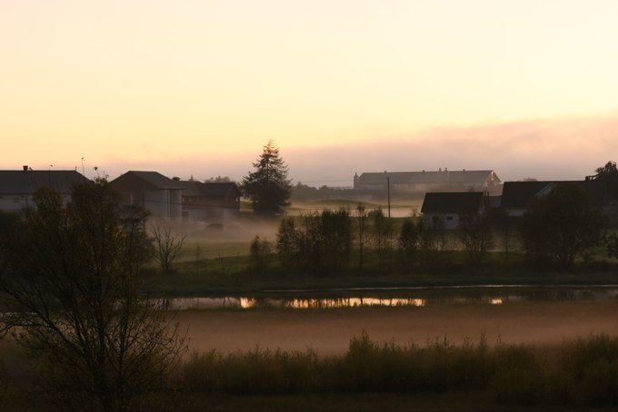 This image captures a serene scene of a small town surrounded by lush green fields at sunrise or sunset, with foggy mountains providing a dramatic backdrop. The calm lake in front adds an element of tranquility to the landscape. In the background, there are several houses and buildings that contribute to the overall charm of the picturesque setting. The colors in the image appear to be particularly vibrant during this time of day, which emphasizes the peaceful atmosphere created by the combination of nature's beauty and human habitation.