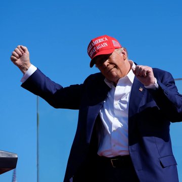 A photo of Donald Trump in a dark suit and red 'Make America Great Again' hat dancing with his arms raised against a clear blue sky.