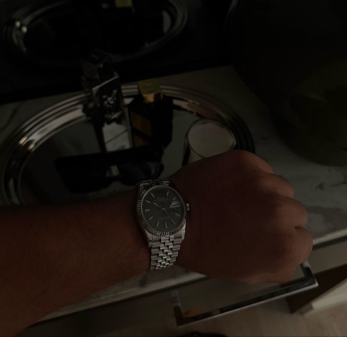 A wrist wearing a silver Rolex watch rests on a reflective surface, with perfume bottles visible in the dimly lit background.