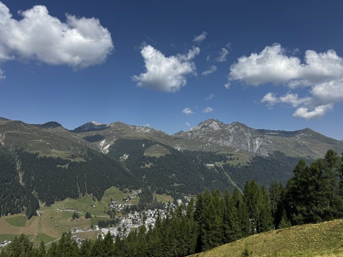 A scenic view of Davos, Switzerland, with its mountain village nestled among lush green alpine slopes under a bright blue sky with scattered clouds. The image complements the post's narrative of the author returning to Davos and humorously claiming to spark a crypto rally with each visit.
