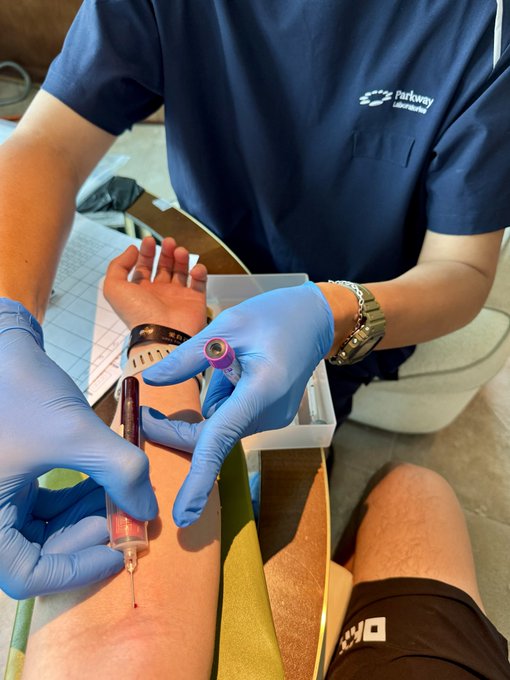 A medical worker wearing blue gloves and a Parkway Laboratories uniform draws blood from a patient's arm into a syringe and collection tube.