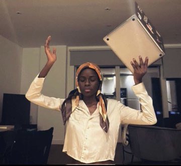 The image features a woman wearing a white shirt, holding a laptop computer above her head with both hands. She has a scarf wrapped around her head and is standing in front of a chair. The background includes a TV screen mounted on the wall behind her. There are also chairs visible in the room. The caption to the image reads "good morning friends of PFP this user isn't an undergraduate anymore watch out".