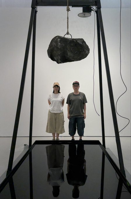 Two people stand inside a large black metal frame beneath a massive suspended boulder in what appears to be an art installation, their reflection visible in a mirror below.
