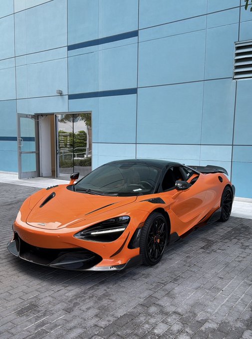 An orange McLaren 720S with a black roof is parked in front of a modern glass building, featuring its signature sleek aerodynamic design and black alloy wheels.