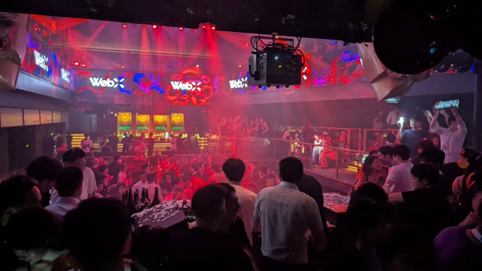 A crowded nightclub scene with red and blue lighting, featuring a stage with screens displaying 'Web', a disco ball, and patrons gathered around the dance floor.