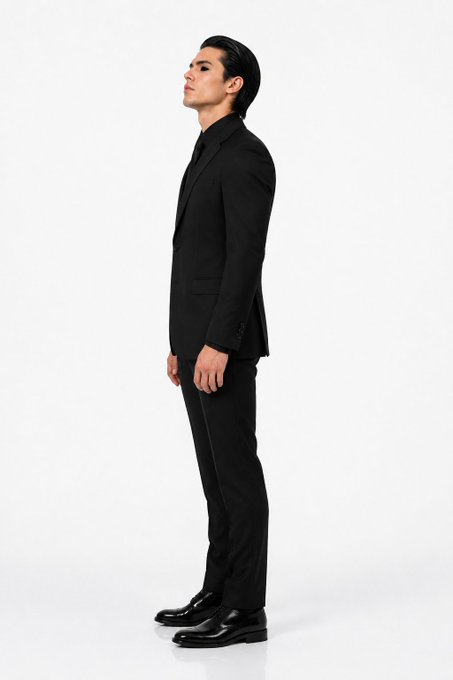 A man with slicked-back dark hair stands in profile, wearing a sleek black suit and black shoes against a light gray background.
