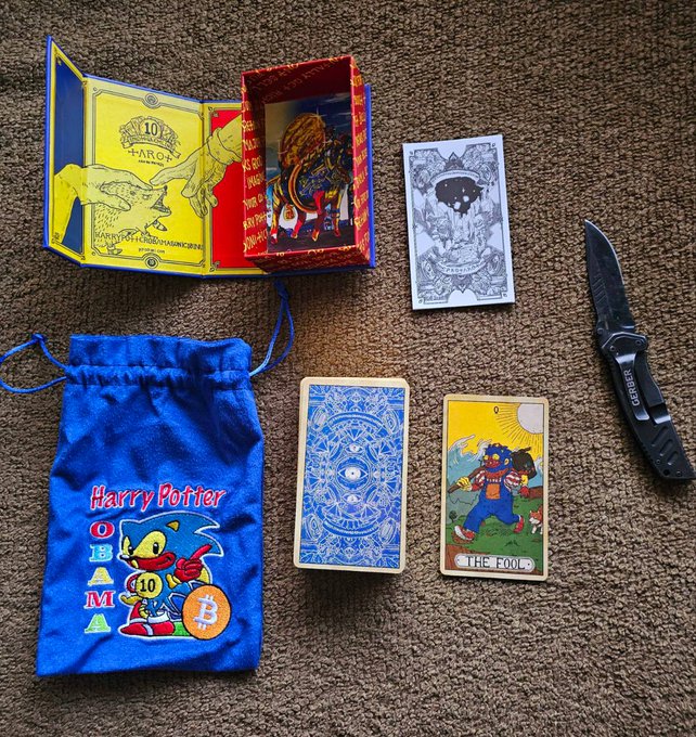 A collection of items on a carpet including a Harry Potter Obama Bitcoin drawstring bag, a 10 Tarot of Joking box containing cards, a 'The Fool' tarot card, and a Gerber pocket knife.