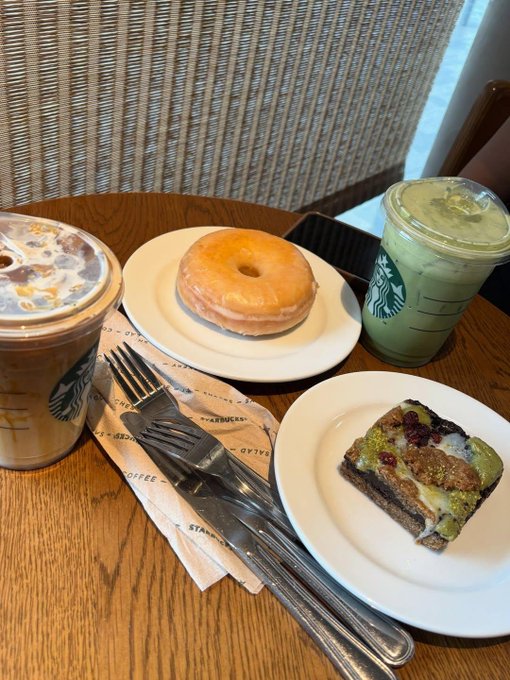 A Starbucks spread on a wooden table features two iced beverages, a glazed donut on a white plate, and a pistachio-topped pastry slice, with cutlery on a Starbucks-branded napkin.