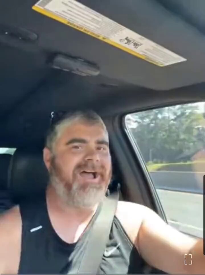 ents, the name of a person with long hair wearing sunglasses, is visible as he drives down the road. He has his mouth open and appears to be having an animated discussion or expressing strong emotions. The car behind him is moving at high speed, which contributes to the sense of excitement and motion in the image. This photograph captures the dynamic nature of a moment where someone's emotions are being articulated through their actions while driving.