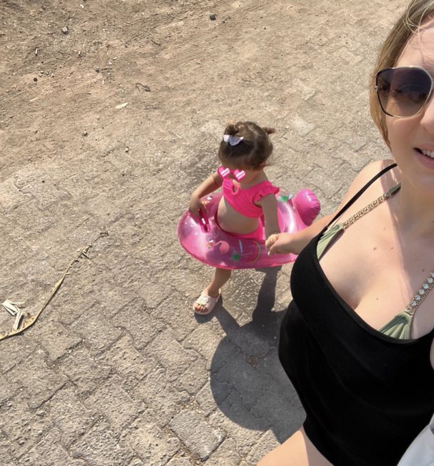 A woman in a black swimsuit and sunglasses takes a selfie while her young daughter, wearing a pink flamingo swimsuit and inflatable ring, walks behind her on a sandy path—perfectly capturing the carefree, sunny "walking into the new week" vibe of the post.