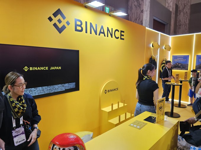 A bright yellow Binance Japan exhibition booth is shown with staff members and visitors. The booth features the 'BINANCE' logo, 'BINANCE JAPAN' signage on a digital screen, and Binance-branded merchandise.