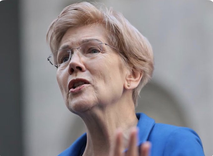 A close-up of an older woman with short blonde hair and glasses wearing a blue blazer, appearing to be speaking.
