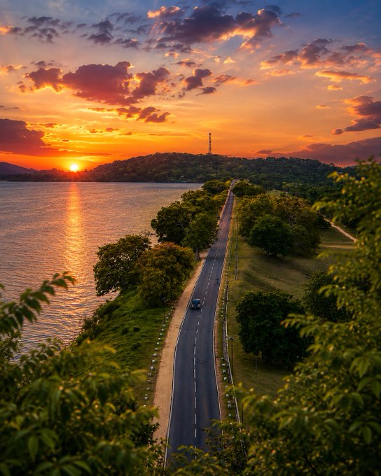 The image shows a beautiful sunset over a river surrounded by trees and hills. A car can be seen driving down a road that runs alongside the water, creating a picturesque scene. The sky is filled with orange and yellow hues as the sun sets behind the horizon. The photo captures the essence of a serene and peaceful moment in nature.