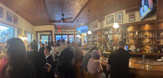 A crowded bar scene with patrons gathered at a wooden bar, featuring a ceiling fan, framed pictures on the walls, and an extensive collection of liquor bottles on shelves behind the bar.