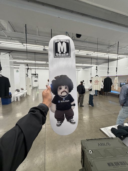 A person holds up a white skateboard deck featuring a 'NEW WORLD ORDER' logo and a cartoon character with an afro wearing a shirt that says 'SOLANA', with a green crate labeled 'ARK/B' visible below.
