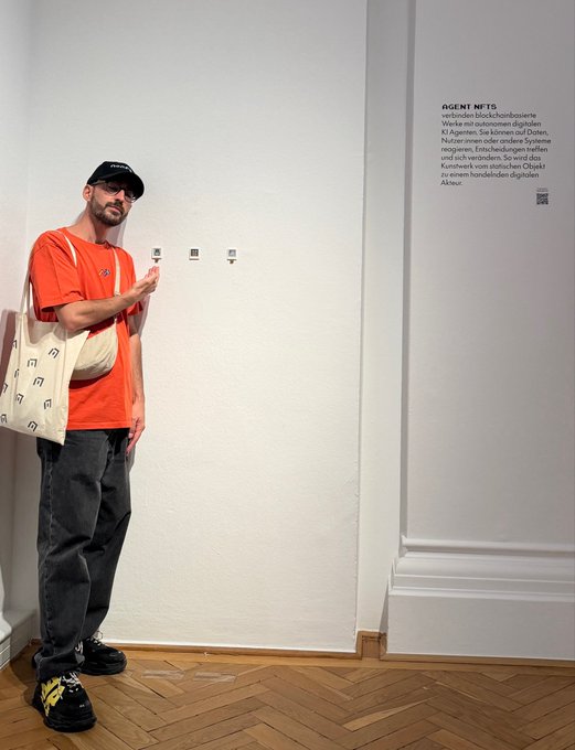 A man wearing an orange shirt, black cap, and carrying a tote bag stands in a gallery, pointing at three small QR codes mounted on a white wall next to a German-language description for 'AGENT NFTS' by Slinky Studios.