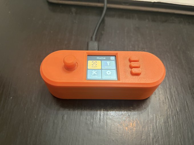An orange 3D-printed handheld device with a small color screen displaying a "Home" menu with icons for "Scan," "Seeds," "Tools," and "Settings," featuring a joystick and three side buttons, sits on a dark wooden surface. The "Kicking it old school" caption references the nostalgic, retro-gaming aesthetic of this DIY Bitcoin hardware wallet prototype.