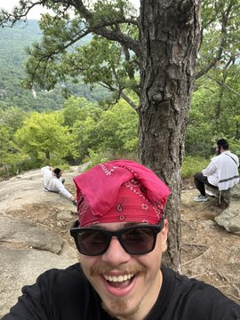 A bearded man in a red bandana and sunglasses smiles at the camera while taking a selfie on a scenic mountain overlook, with friends sitting on rocks in the background. The post celebrates reaching the "peak" of their hike with Jewish friends, using the hiking summit as a playful metaphor to request USDT cryptocurrency donations.