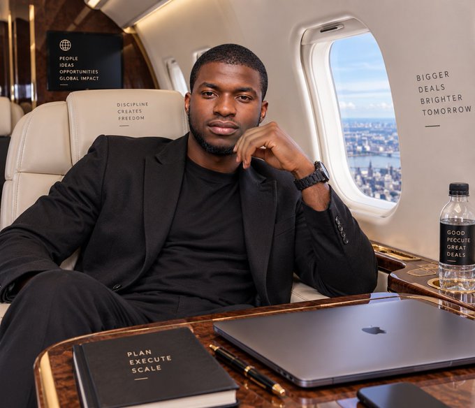 A man in a black suit and t-shirt sits inside a private jet, posing thoughtfully near a laptop and a notebook labeled 'PLAN EXECUTE SCALE', with motivational text on the surrounding surfaces.
