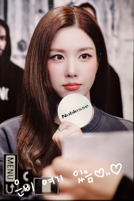 A polaroid-style photo of a young woman with long brown hair holding a round 'Noblesse' compact mirror, with Korean text and heart decorations at the bottom.
