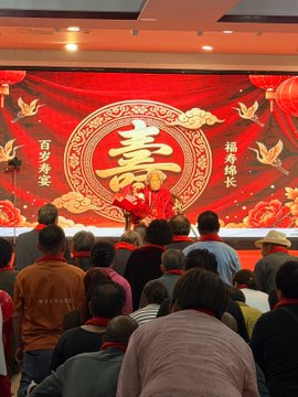 An audience gathers before a large red stage backdrop featuring the Chinese character "寿" (longevity), with the phrases "百岁宴" (Centennial Birthday Banquet) and "福寿绵长" (Enduring Blessings and Longevity) flanking it, celebrating an elderly grandmother's 100th birthday as mentioned in the post.
