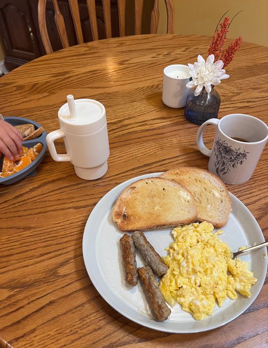 A breakfast plate featuring scrambled eggs, sausage links, and toasted bread sits on a wooden table alongside a coffee mug, a small vase with flowers, and a teapot, capturing a peaceful morning moment that pairs perfectly with the "Blessed" sentiment.