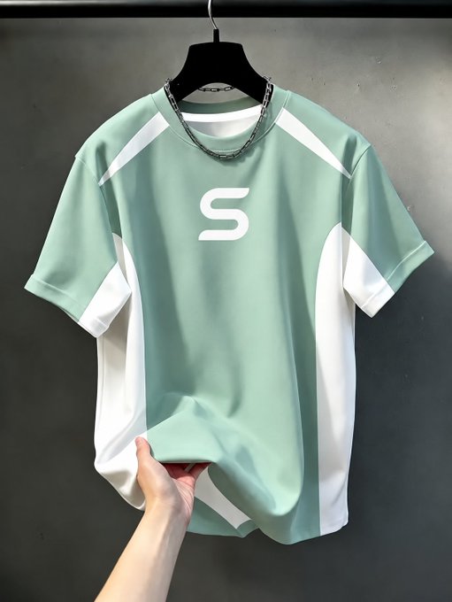 A short-sleeved t-shirt in light green and white is displayed on a black hanger, featuring a prominent 'S' logo on the chest and a silver chain detail on the neckline.