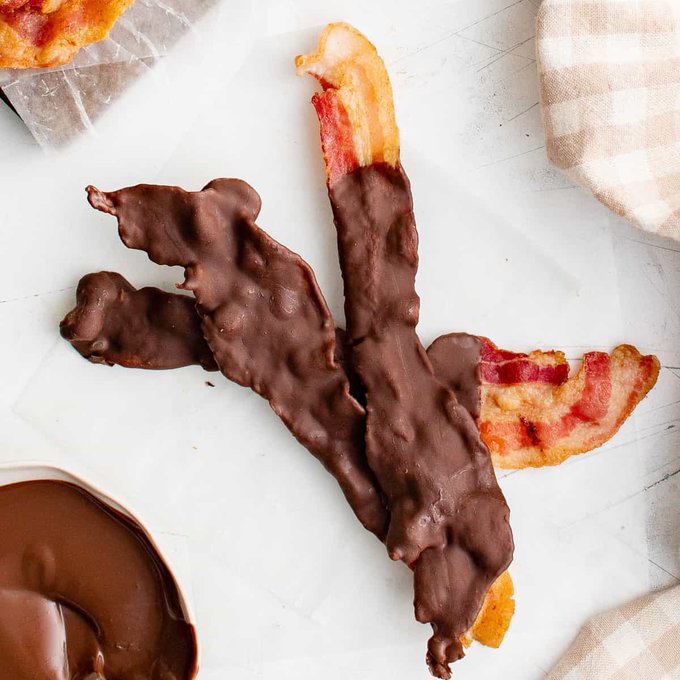 Strips of bacon dipped in melted chocolate arranged on a white surface next to a bowl of chocolate and a checkered cloth.