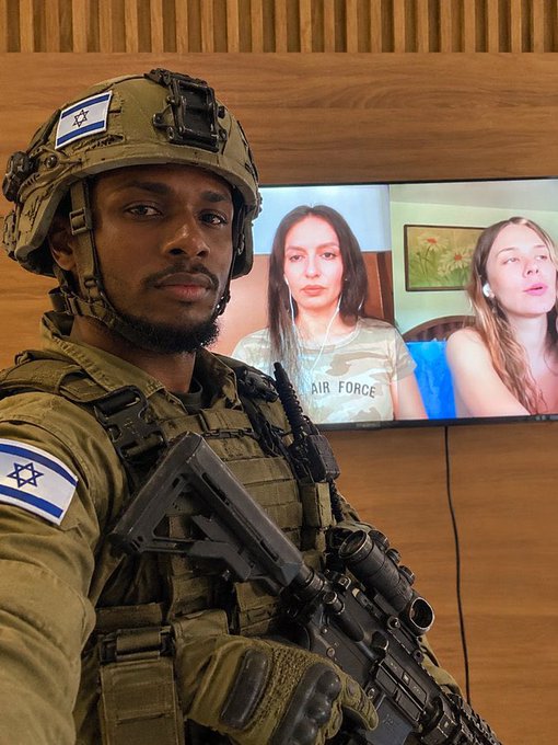 An Israeli soldier in tactical gear with an Israel flag patch on his helmet stands in front of a TV screen displaying two women, one wearing an 'AIR FORCE' shirt, in what appears to be a video call.
