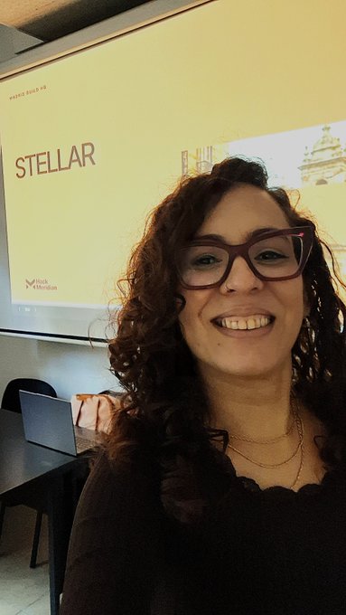 A smiling woman with curly dark hair and red glasses taking a selfie in front of a projection screen displaying 'STELLAR' and the Merk Mokum logo.