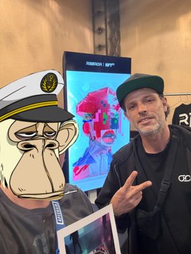 A man wearing a dark cap and jacket stands next to a cartoon monkey cutout wearing a captain's hat, with a digital display showing colorful artwork behind them.