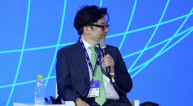 A man in a dark suit and green tie speaks into a microphone at what appears to be a conference or panel discussion, with a blue geometric-patterned backdrop behind him and a water bottle visible on a nearby table. The image, showing a speaker at what is likely a BitMine-related corporate or crypto industry event, accompanies the post announcing BitMine's massive Ethereum holdings and its addition to the Russell 1000 index.
