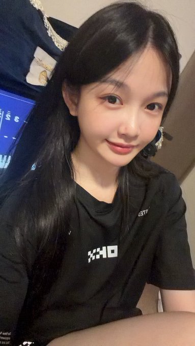 A selfie of a young woman with long black hair wearing a black shirt with white text, indoors with a computer monitor in the background.