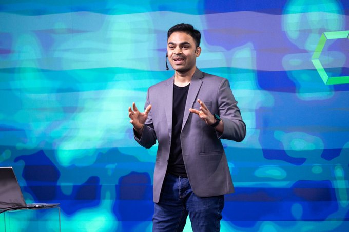A man in a gray blazer over a black t-shirt gives a presentation on a stage with a bright blue and teal patterned background, gesturing with both hands while wearing a small headset microphone.